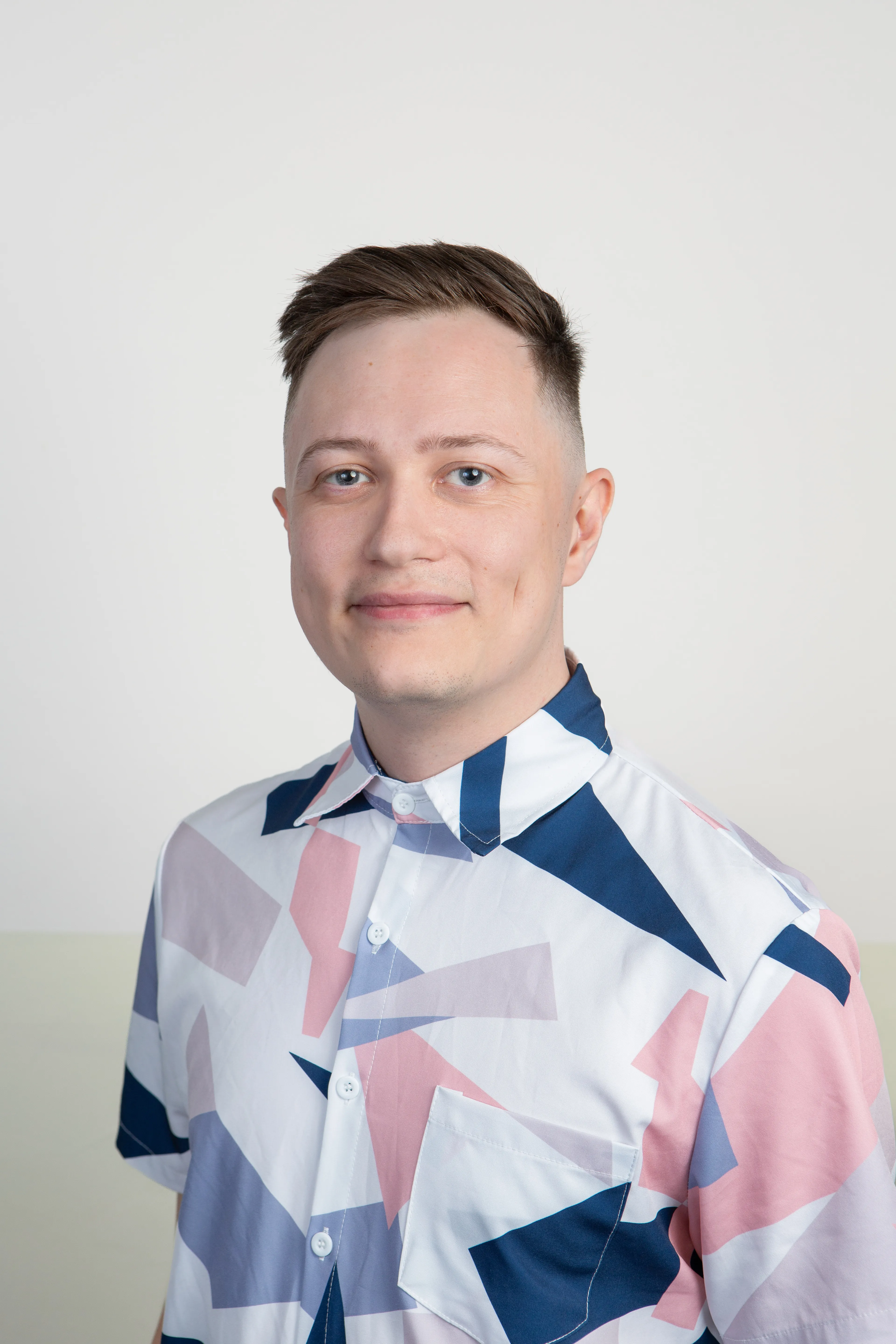 A studio headshot of Chris Parsons, a person with short, styled brown hair and blue eyes, smiling gently at the camera. They are wearing a white, short-sleeved button-down shirt featuring a bold geometric pattern in shades of blue, pink, and lavender. The background is a clean, minimalist split of off-white on top and a soft, muted sage green on the bottom.